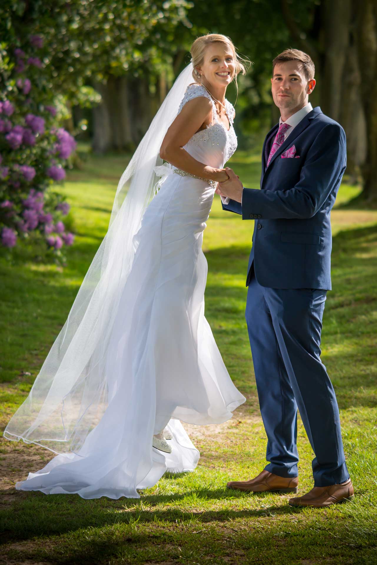 Wedding couple, she is wearing her dress and veil, smiling towards camera, shown jumping, holding his hands, she is a PE teacher.