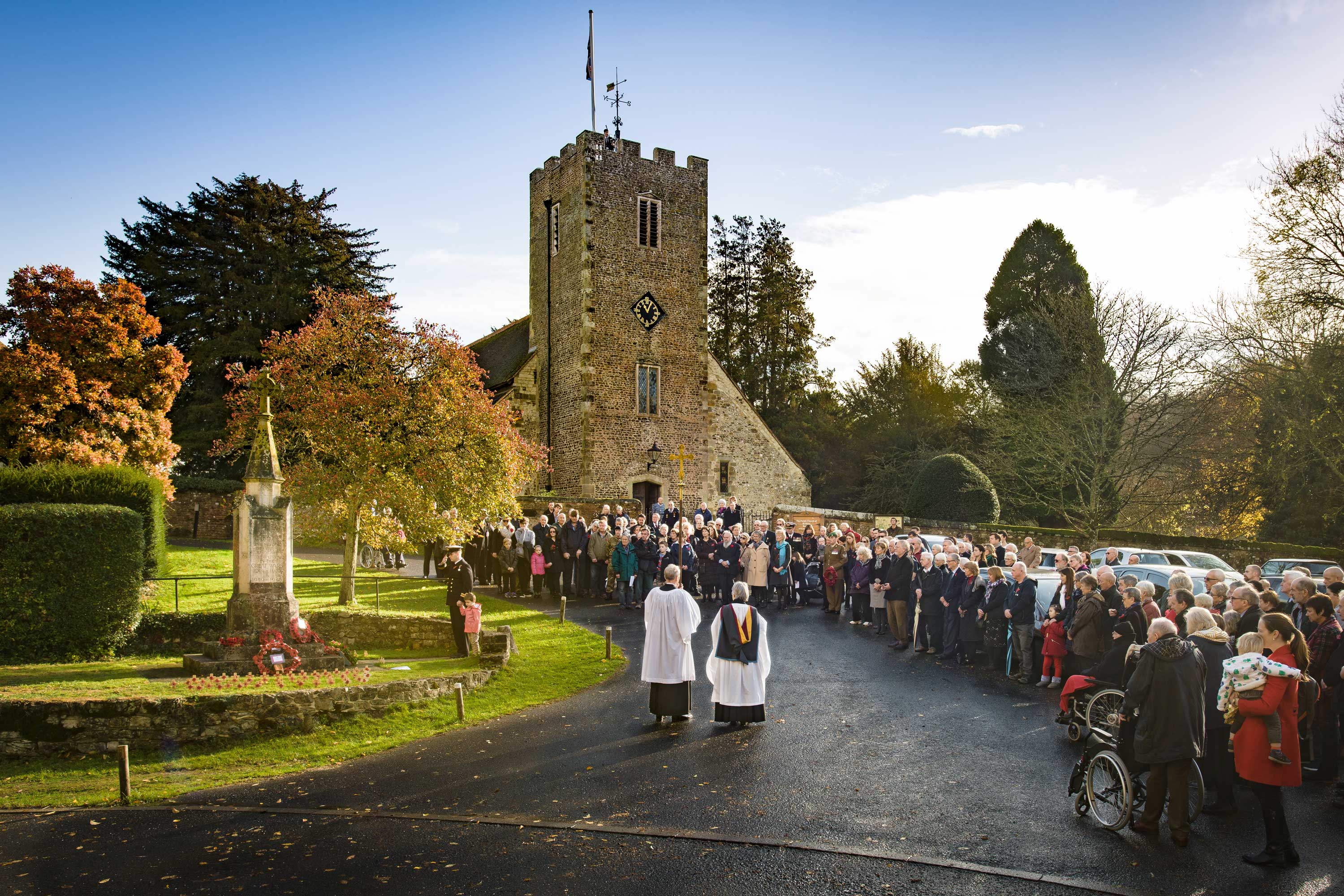 View of the salute to the war memorial at rememberence day, many people are gathered round and the piper is at the top of the church tower ready to play the lament 'Flowers of the Forest'.  The church clock shows two minutes past eleven.
