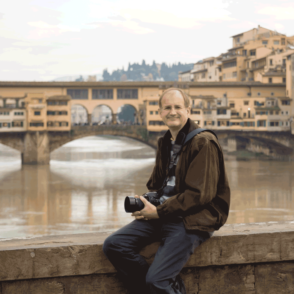 Jon holding a camera, sat on a bridge parapet in Florence, Italy, looking towards the taker, the Ponte Vecchio (old medieval stone bridge) over the river Arno is in the background.