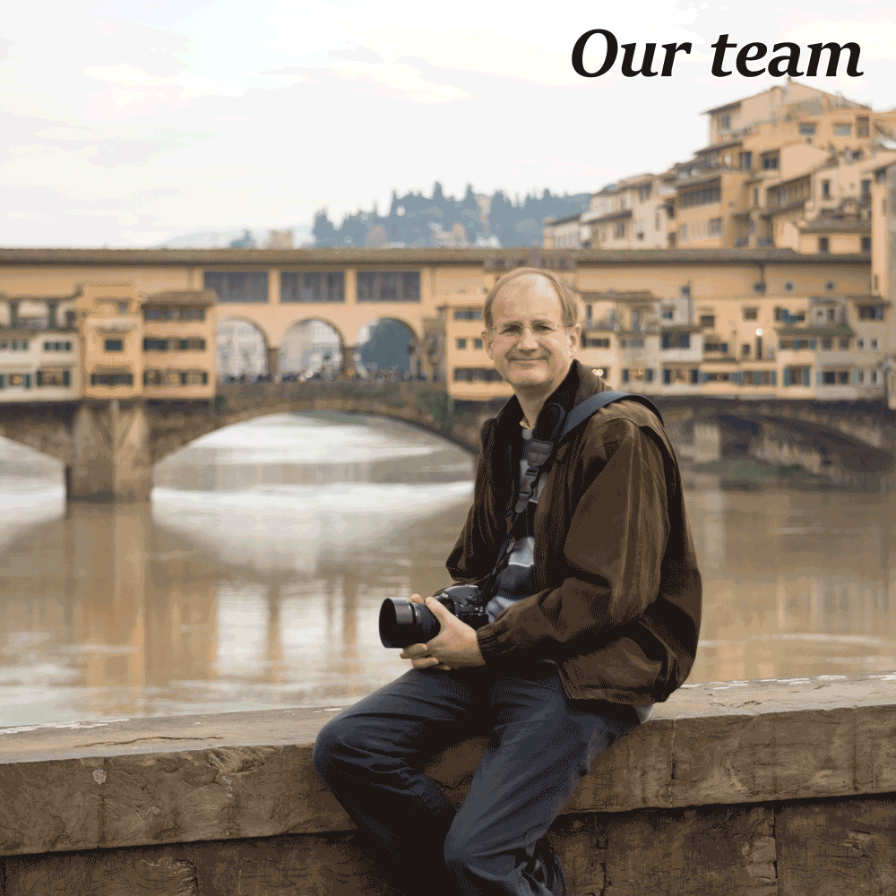 One of our team holding a camera, sat on a bridge parapet in Florence, Italy, looking towards the taker, the Ponte Vecchio (old medieval stone bridge) over the river Arno is in the background.