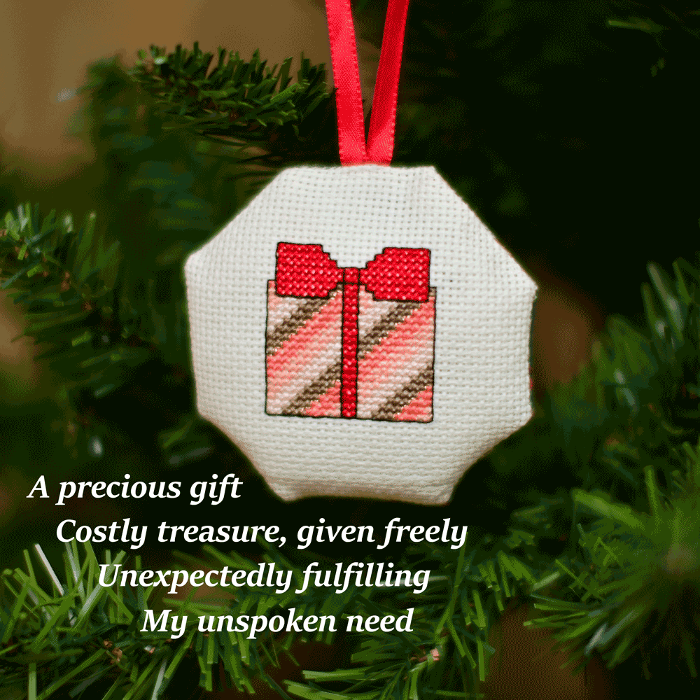 Cross-stitched octagonal picture of a present, hanging in a Christmas tree.