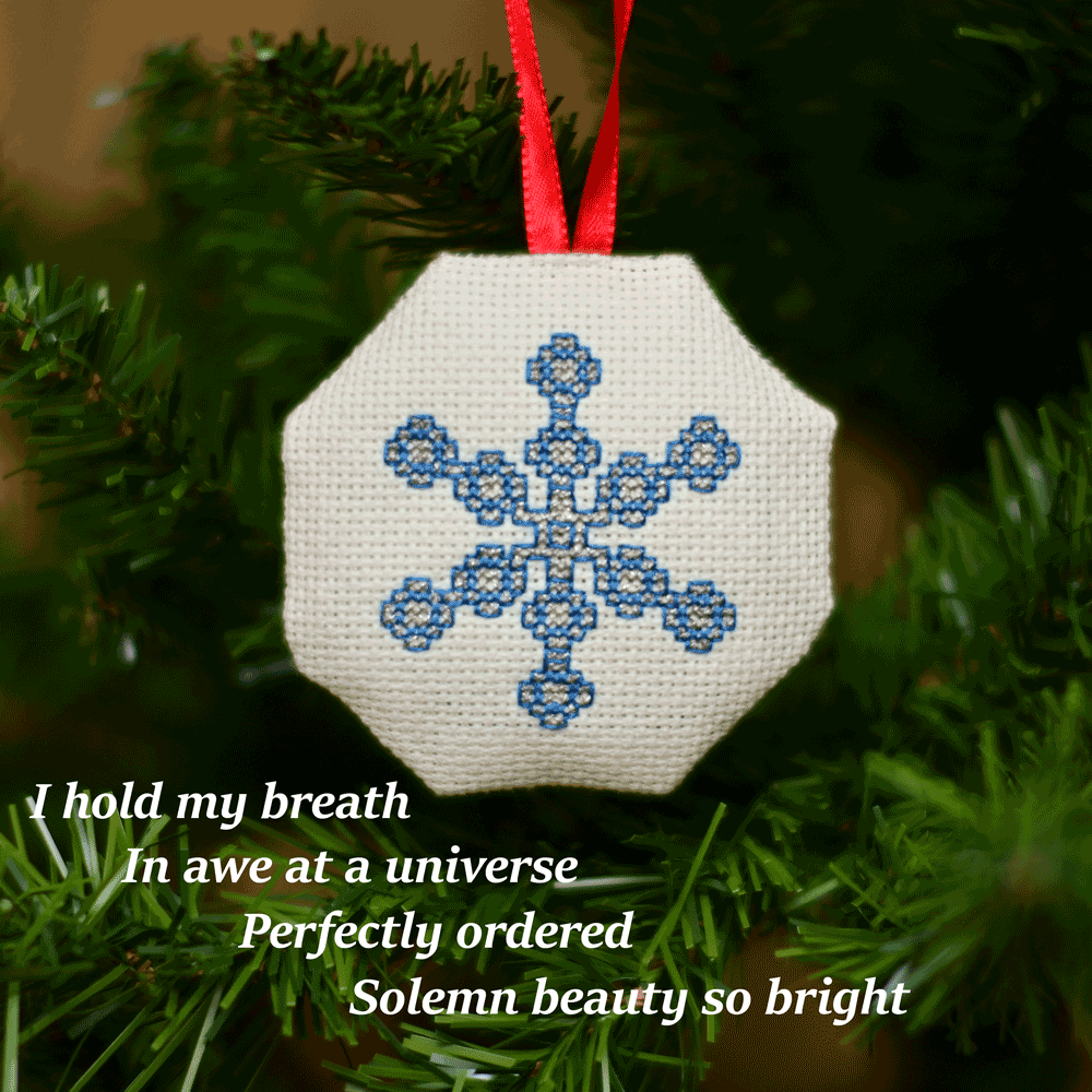 Cross-stitched octagonal picture of a snowflake, hanging in a Christmas tree.