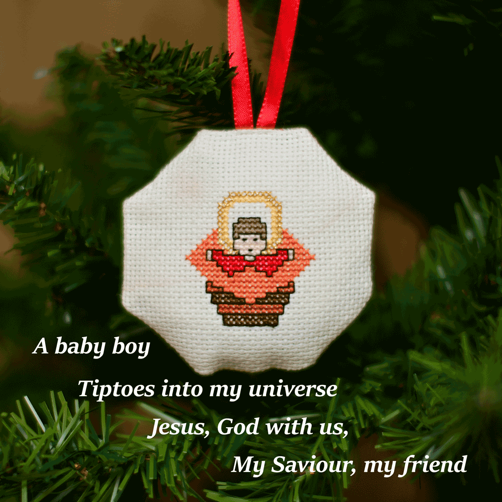 Cross-stitched octagonal picture of the baby Jesus in the manger, hanging in a Christmas tree.